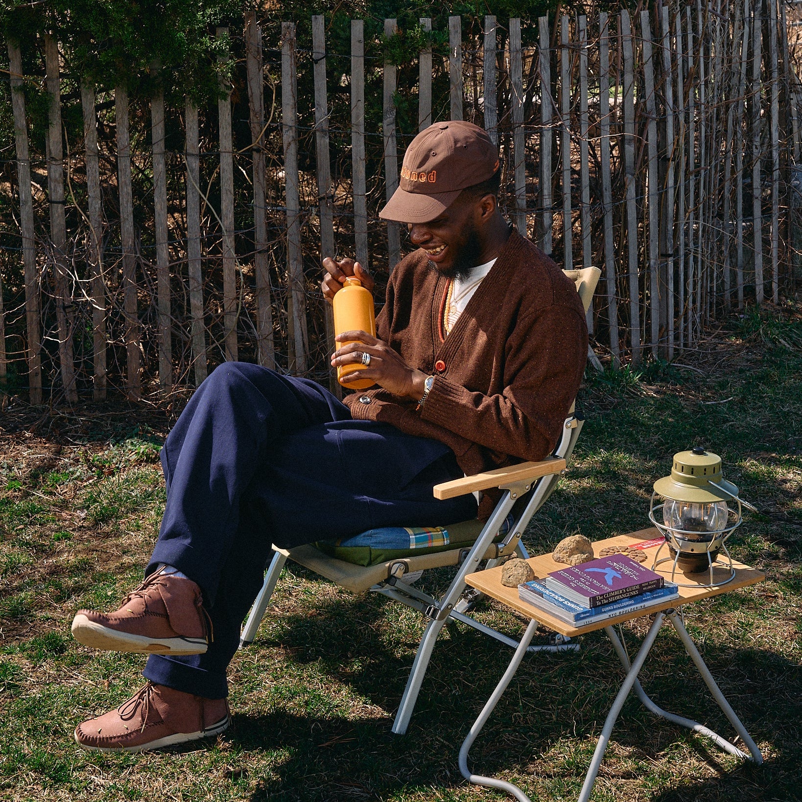 Man sitting outdoors with a small table, holding a bottle, wearing a cap and brown sweater.